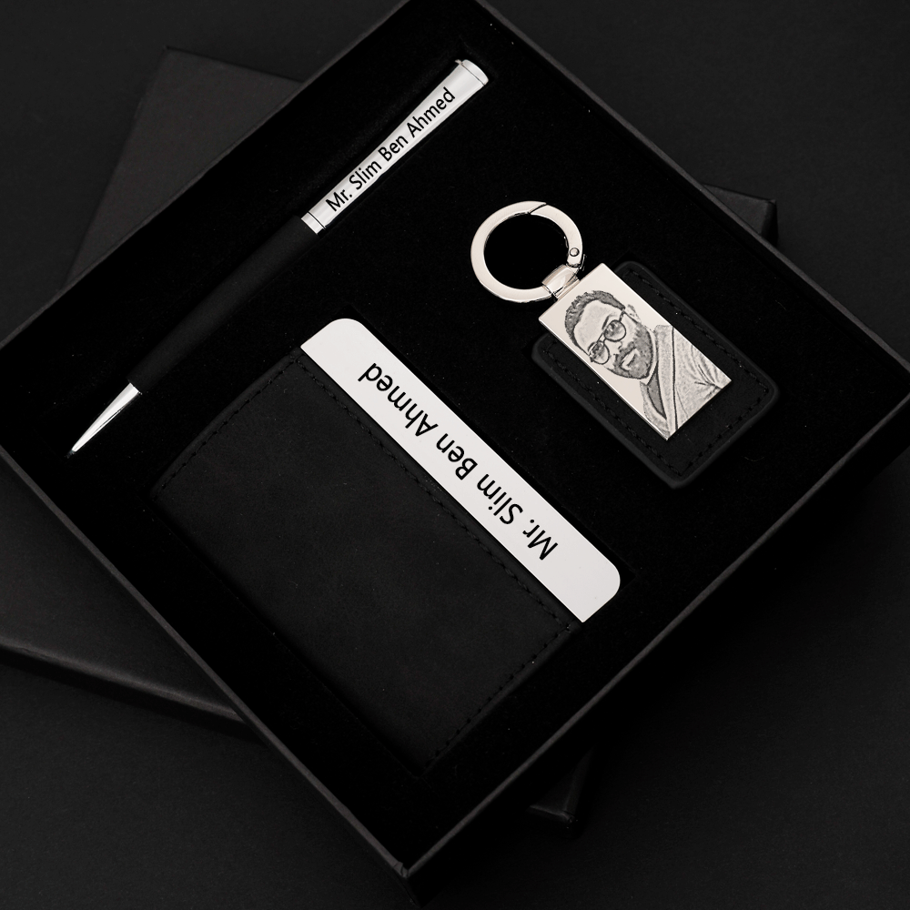 Un coffret cadeau noir contenant un stylo, un porte-clés et un portefeuille. Le stylo est noir avec une partie argentée et porte l'inscription \"Mr. Slim Ben Ahmed\". Le porte-clés est en métal avec une gravure d'un visage et est attaché à une étiquette en cuir noir. Le portefeuille est noir et contient une carte blanche avec le nom \"Mr. Slim Ben Ahmed\" écrit dessus.